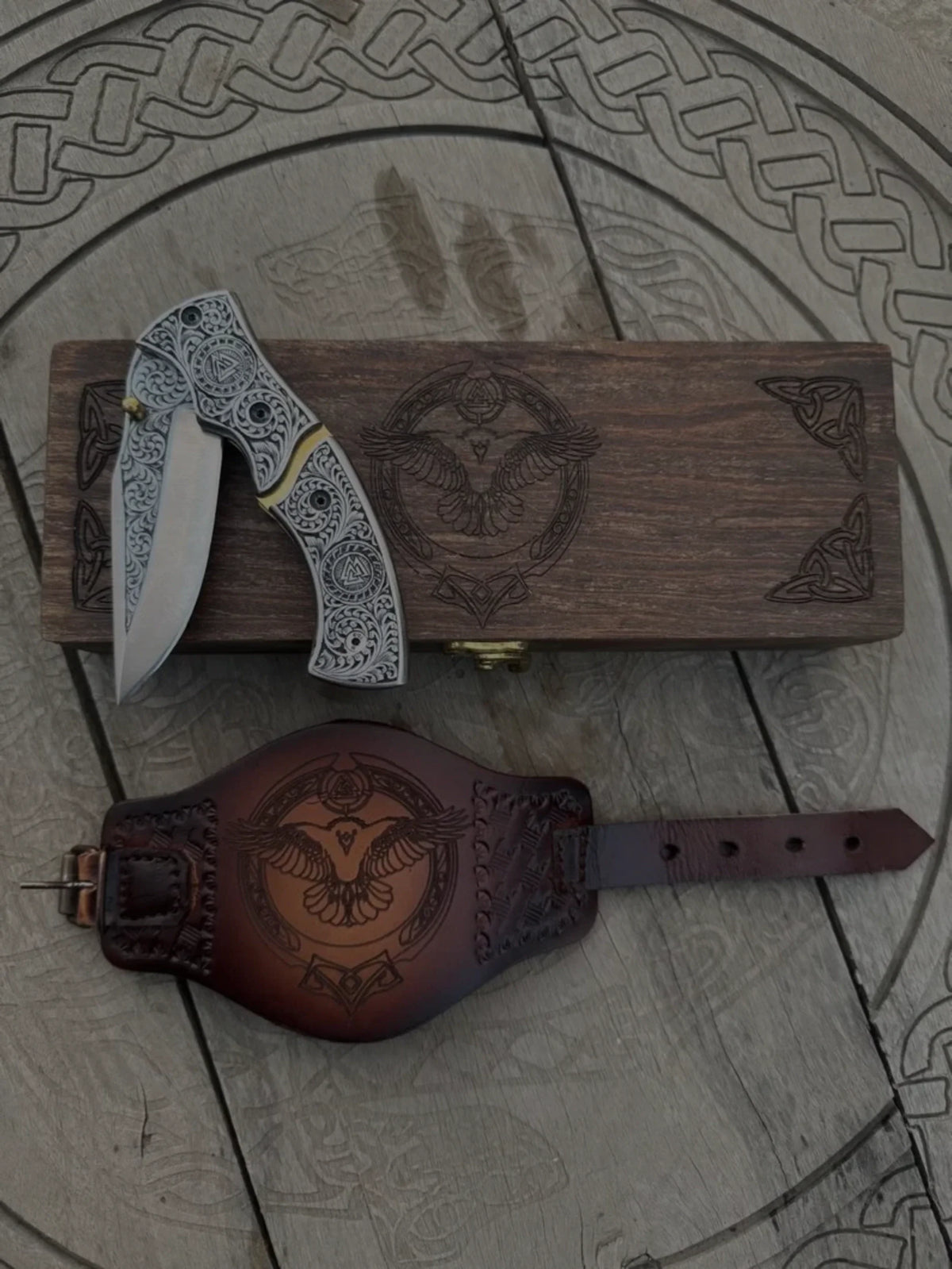 Engraved Viking folding pocket knife with Norse Valknut patterns, displayed with a matching carved wooden gift box and an embossed brown leather sheath featuring a raven design
