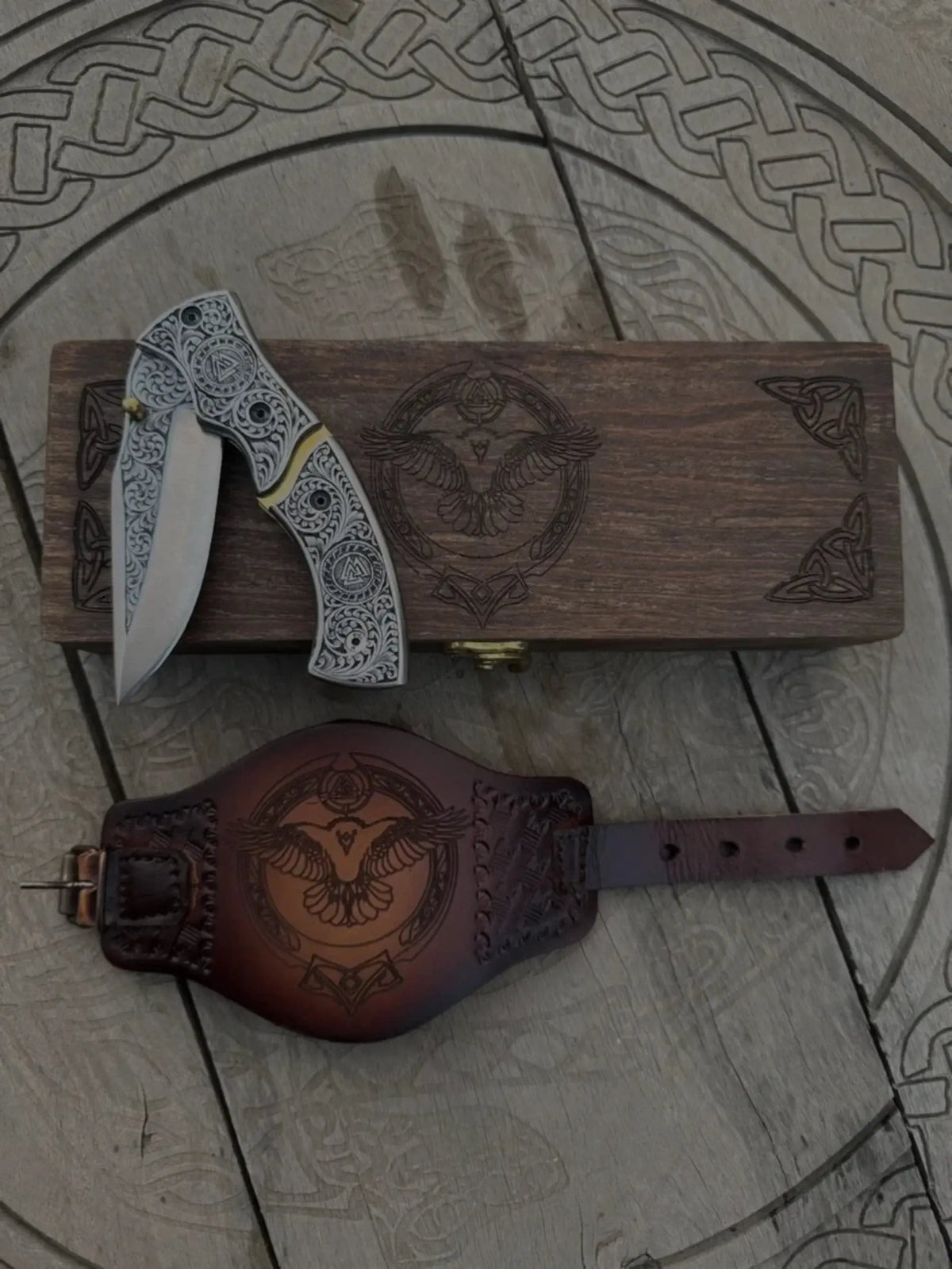 Engraved Viking folding pocket knife with Norse Valknut patterns, displayed with a matching carved wooden gift box and an embossed brown leather sheath featuring a raven design