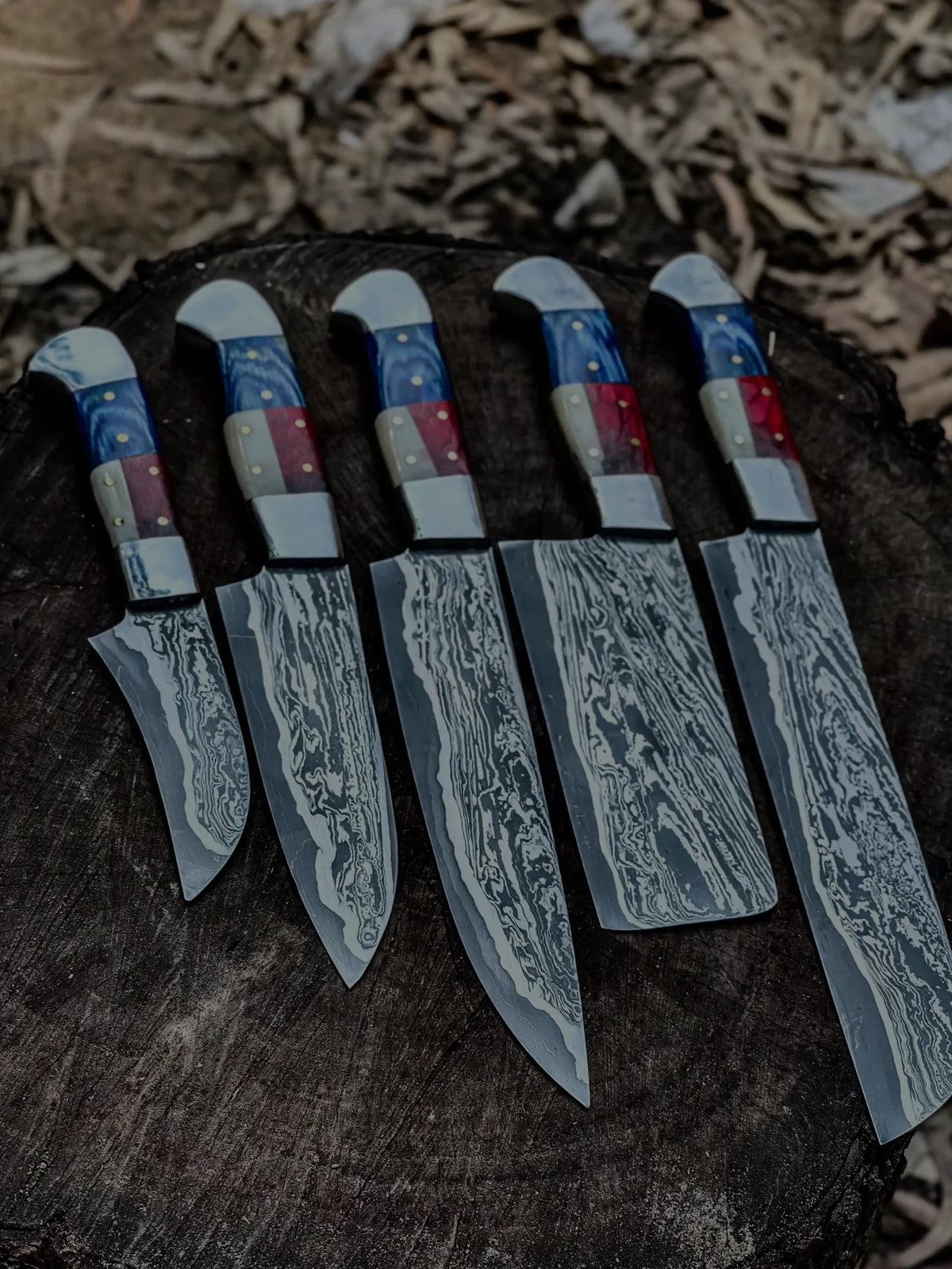 A 5-piece custom Damascus steel chef knife set with unique blue, red, and white multi-colored handles, displayed vertically on a rustic tree stump background