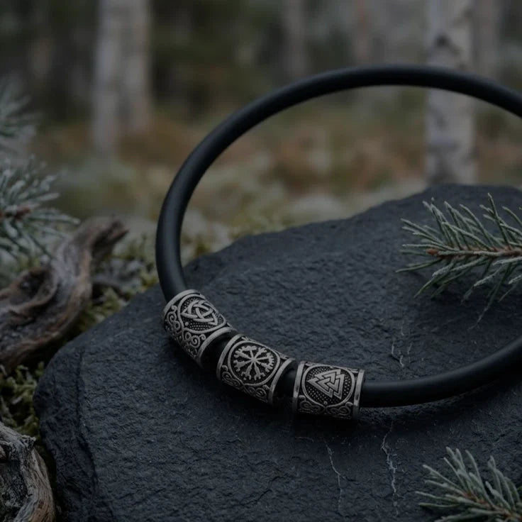 Handcrafted black cord Viking torc necklace featuring silver-toned beads engraved with a Triquetra, Vegvisir, and Valknut symbol, resting on a dark slate stone outdoors.