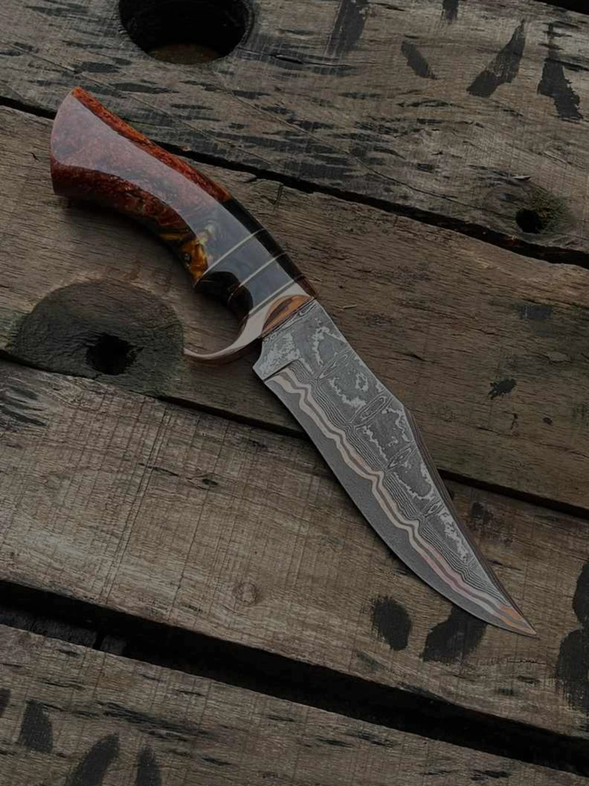 Custom hand-forged Damascus steel hunting knife with a distinct copper layer pattern on the blade, featuring a polished burl wood and red resin hybrid handle, resting on a rustic wooden pallet