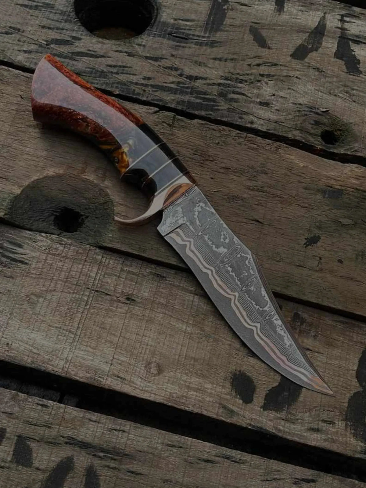 Custom hand-forged Damascus steel hunting knife with a distinct copper layer pattern on the blade, featuring a polished burl wood and red resin hybrid handle, resting on a rustic wooden pallet