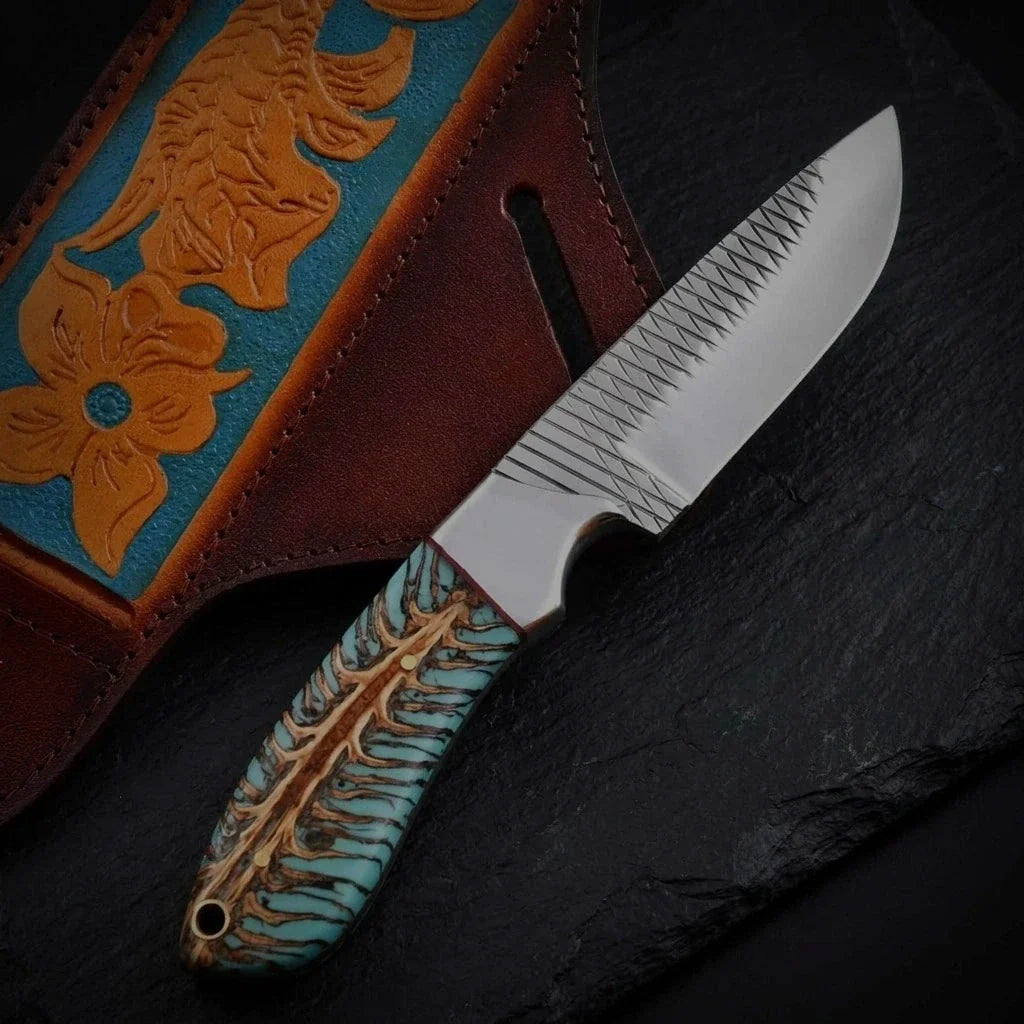 Handcrafted fixed-blade hunting knife featuring a turquoise pinecone resin handle and cross-hatched blade spine, resting beside a tooling-carved leather sheath on a dark stone surface