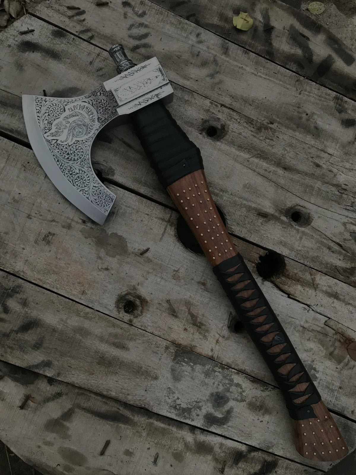 Hand-forged Viking axe with an intricately engraved steel blade, detailed Nordic patterns, and a wooden handle wrapped in black leather, resting on a rustic wooden surface