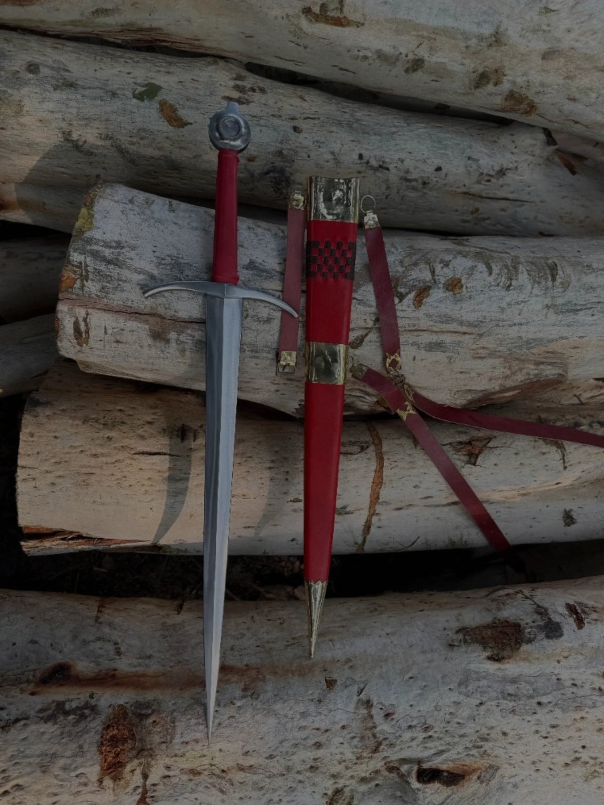 Handcrafted medieval sword with a red leather-wrapped hilt alongside a matching red leather scabbard with brass fittings, resting on rustic wooden logs