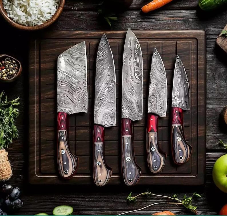 5-piece Damascus steel kitchen knife set featuring ergonomic red-and-brown resin wood handles, laid out on a dark wooden cutting board surrounded by fresh vegetables.