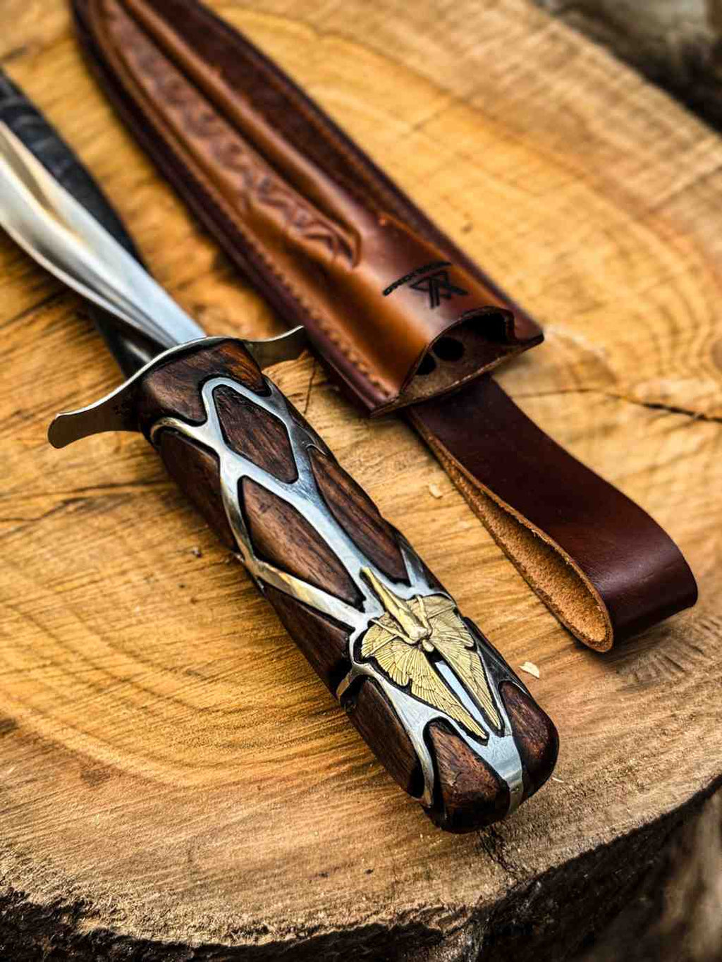 Angled view of a handcrafted Subtle Knife replica with a detailed metal inlay wooden handle and silver blade hilt, displayed next to its brown leather sheath on a rustic log for USA fantasy collectors.