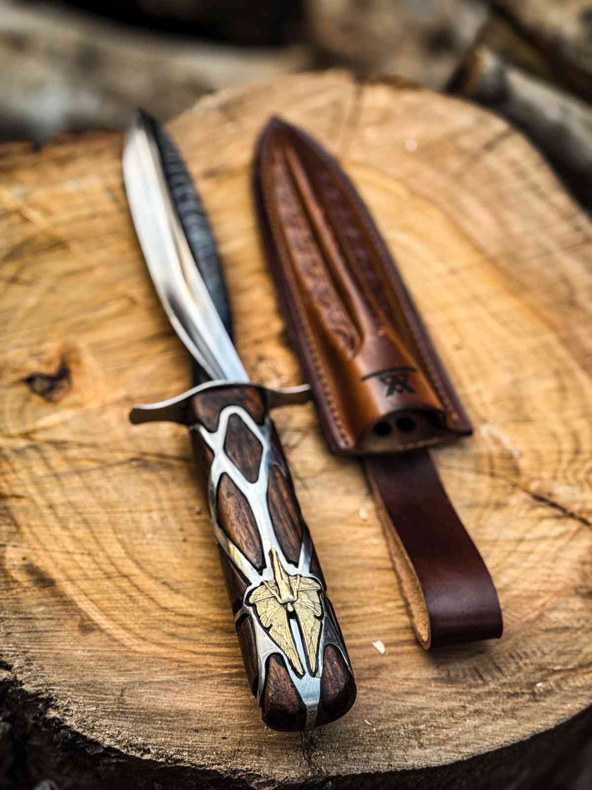 Close-up of a custom-made Subtle Knife replica handle featuring intricate metalwork inlay and wood, resting on a log next to an artisan leather scabbard, handmade for fantasy collectors in the USA.