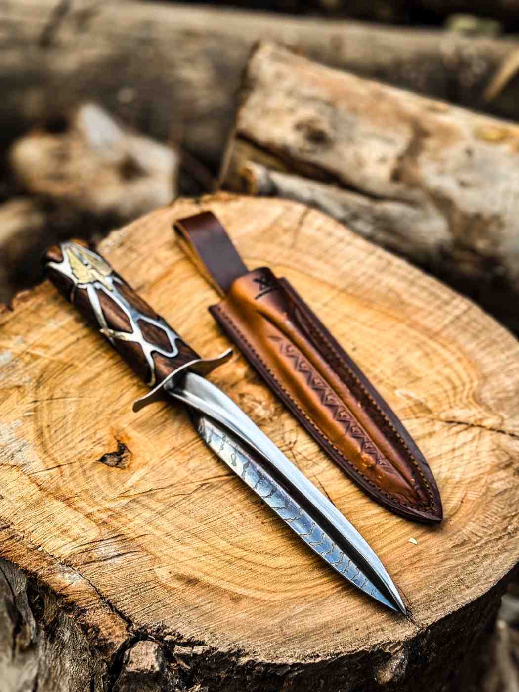 Full view of a handcrafted Subtle Knife replica showing the detailed dual-tone metallic blade and intricate wood handle, lying next to a brown leather sheath on a wood stump for USA fantasy collectors.