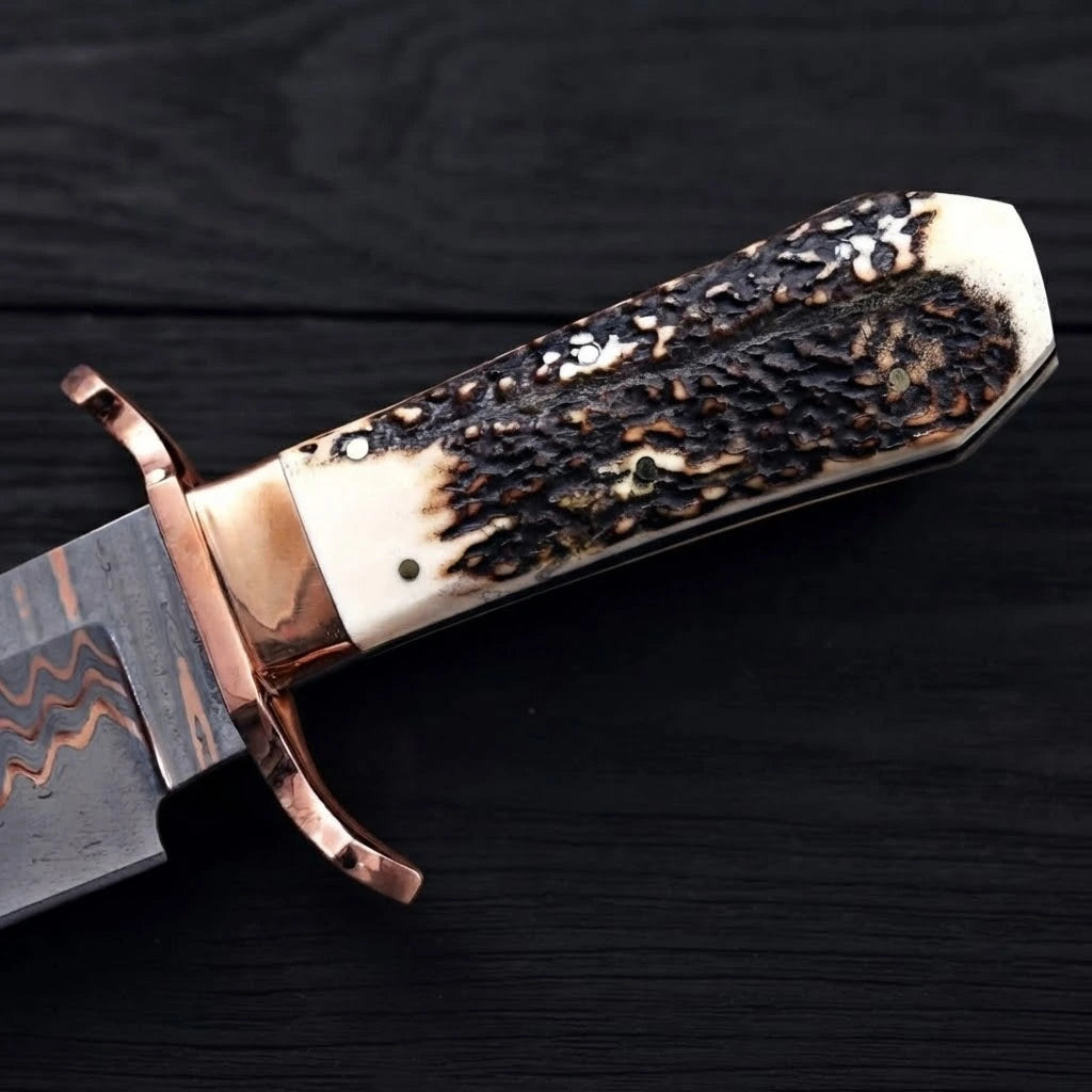 Close-up view of a Bowie knife handle showing a polished copper double guard and a highly textured stag antler scale grip on a dark background