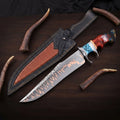 Copper Damascus Bowie knife with a steel double guard, turquoise and red resin hybrid handle, next to a black and brown leather sheath on a dark wood background.