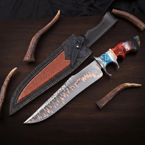Copper Damascus Bowie knife with a steel double guard, turquoise and red resin hybrid handle, next to a black and brown leather sheath on a dark wood background.