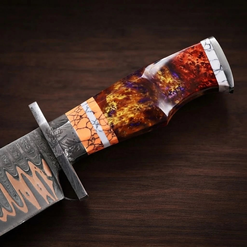 A detailed close-up of a custom Bowie knife handle, showcasing a polished fire resin grip with rich red, orange, and gold tones, accented by orange and white marbled stone spacers, a steel handguard with engraved scrollwork, and a partial view of the copper Damascus blade, on a dark wooden background
