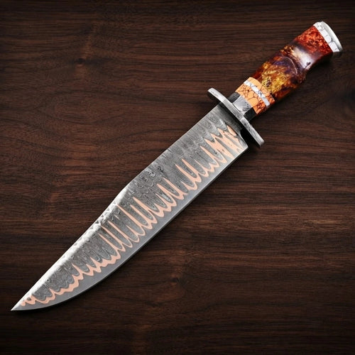 A full diagonal profile view of a custom Copper Damascus steel Bowie knife, showcasing its distinctive layered wavy blade pattern with copper tones, a steel handguard, and a vibrant orange-red fire resin and faux stag hybrid handle, resting on a dark walnut wood grain background