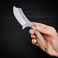 A hand holding the Cowboy Cutter Knife, showcasing the cross-hatched rasp steel blade texture and natural wood handle against a dark wood background