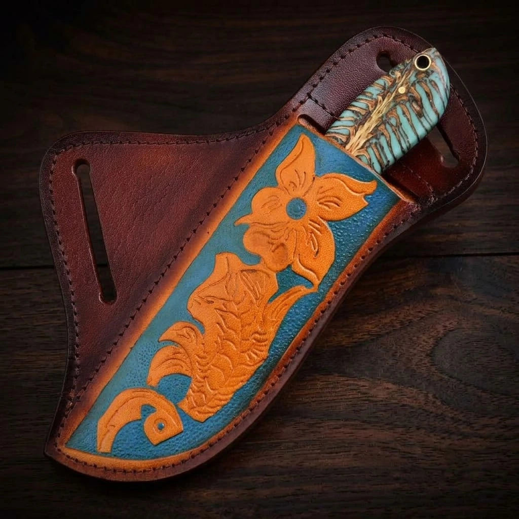 A handmade USA cowboy knife securely sheathed inside its custom brown leather crossdraw holster, showing only the turquoise-dyed pine cone hybrid handle, with the sheath displaying vibrant orange floral carvings on a blue background over a dark wooden surface.