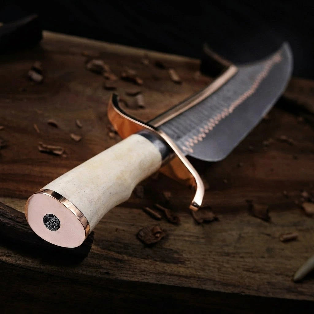 A selective focus, angled view of a custom handmade Damascus Bowie knife resting on a rustic wood surface, highlighting the polished white bone handle, smooth brass pommel cap with a circular inlay, and the curved brass handguard extending toward the blade.