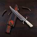 A custom hand-forged Bowie knife featuring a serrated or layered Damascus steel blade with a brass spine and guard, paired with a polished stag bone handle, resting diagonally over a multi-toned brown and black textured leather sheath surrounded by small antler pieces on a dark wooden background