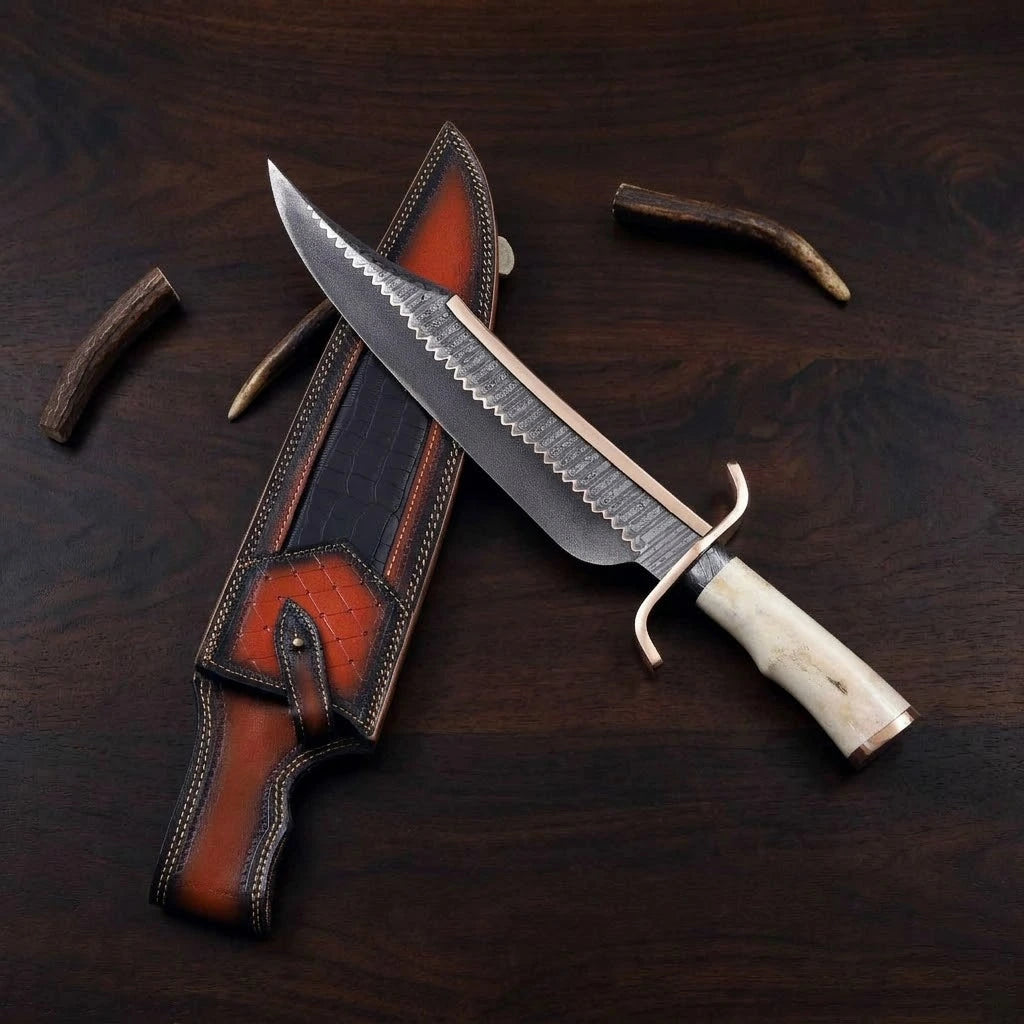 A custom hand-forged Bowie knife featuring a serrated or layered Damascus steel blade with a brass spine and guard, paired with a polished stag bone handle, resting diagonally over a multi-toned brown and black textured leather sheath surrounded by small antler pieces on a dark wooden background