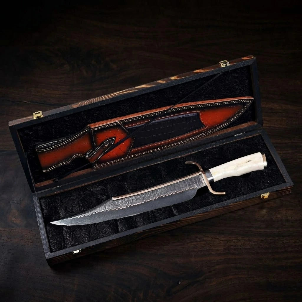A custom hand-forged Damascus steel Bowie knife with a polished bone handle and brass guard, displayed inside an open dark wood presentation gift box with black velvet lining, sitting alongside its multi-textured leather sheath on a dark wooden surface.