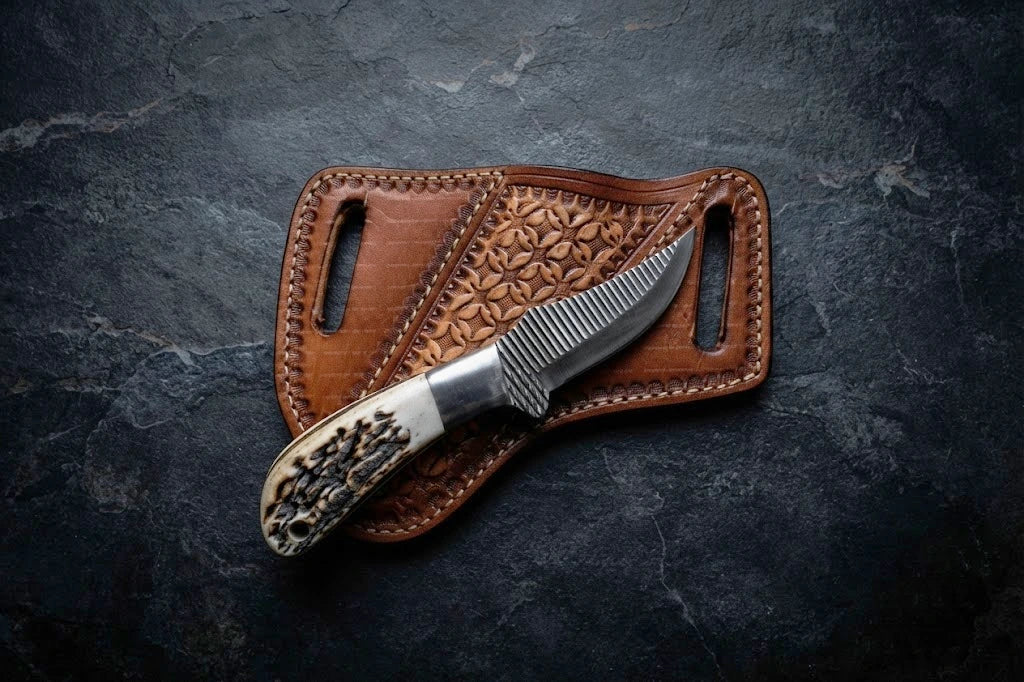 A top-down view of a custom handmade cowboy belt knife forged from a steel rasp file, showing distinct horizontal ridge lines on the blade, a polished metal bolster, and a textured stag antler horn handle, resting on top of a brown floral-tooled leather pancake sheath over a dark slate stone background