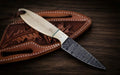 A handmade Damascus steel cowboy belt knife featuring a patterned drop-point blade, a brass bolster with decorative star pins, and a polished bone handle divided by a thin turquoise accent spacer, resting on a brown leather pancake sheath tooled with oak leaf patterns on a dark wooden background