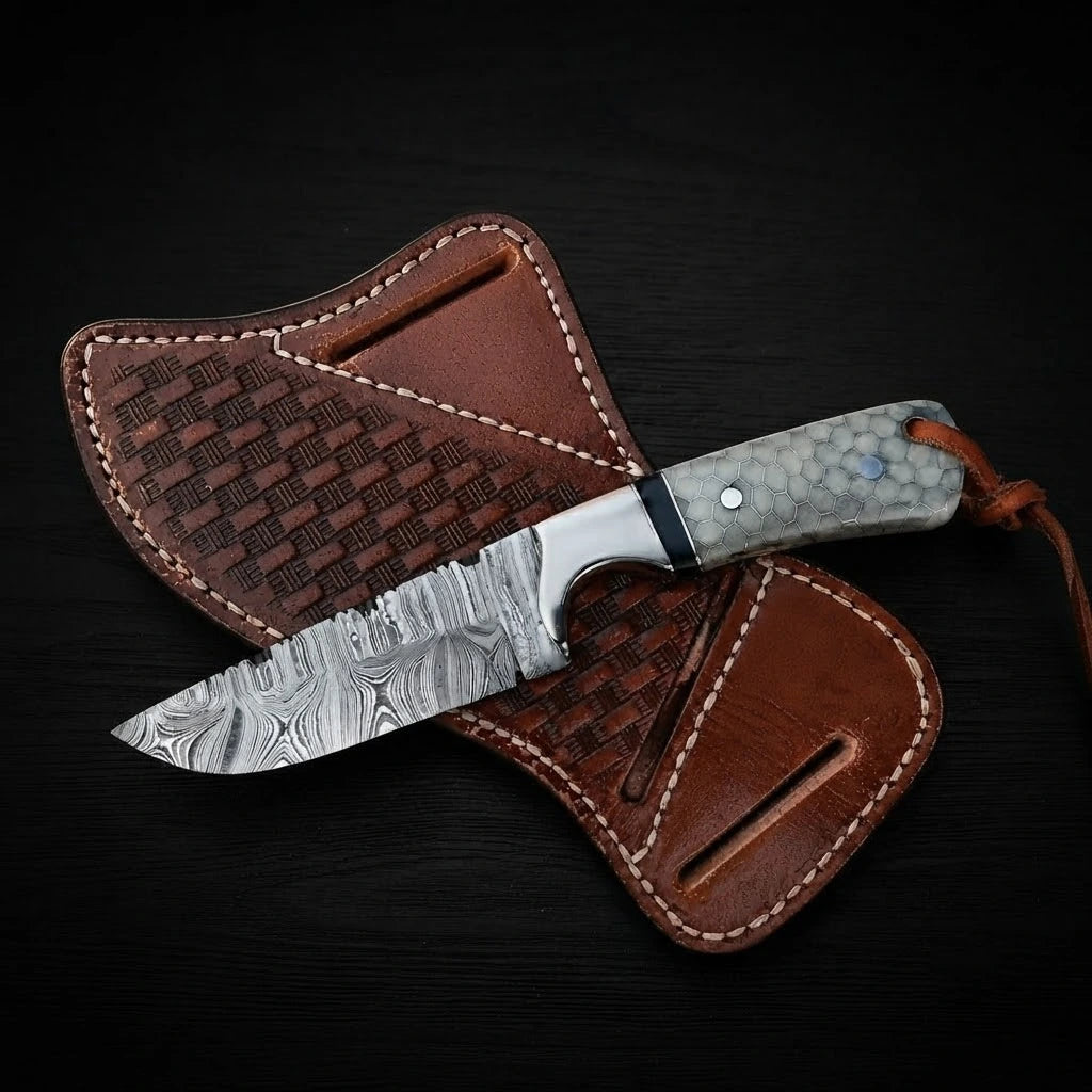 Damascus steel Buckaroo ranch knife with a honeycomb pattern resin handle and leather lanyard, resting on a brown basket-weave leather pancake sheath over dark wood.