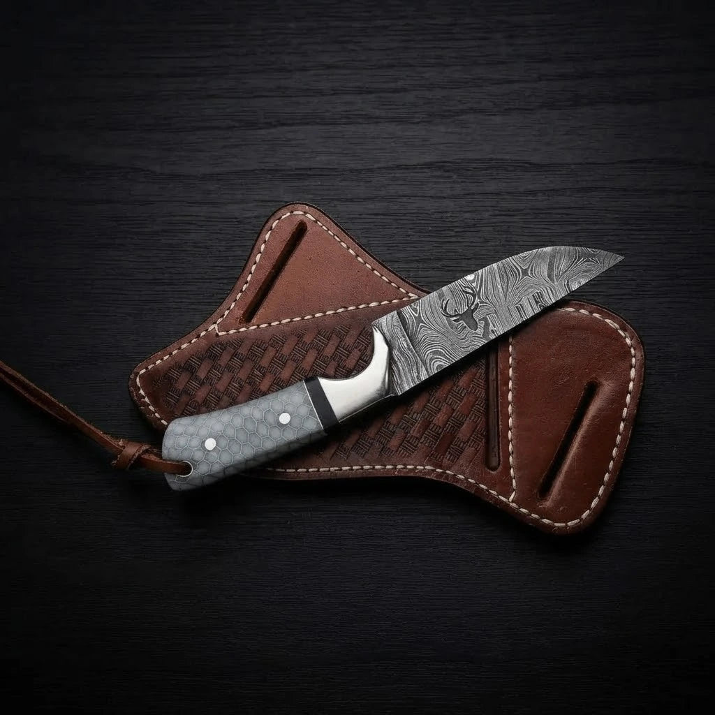 Damascus steel Buckaroo ranch knife featuring a stag logo etching on the blade, honeycomb pattern handle, and leather lanyard, resting on a brown leather pancake sheath over dark wood