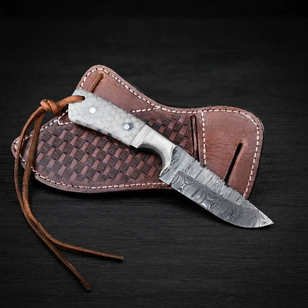 Top-down view of a Damascus steel Buckaroo ranch knife with a honeycomb resin handle and leather lanyard cord, resting on a stamped brown leather pancake sheath over dark wooden background.