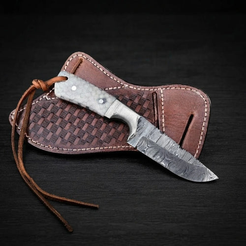Top-down view of a Damascus steel Buckaroo ranch knife with a honeycomb resin handle and leather lanyard cord, resting on a stamped brown leather pancake sheath over dark wooden background.