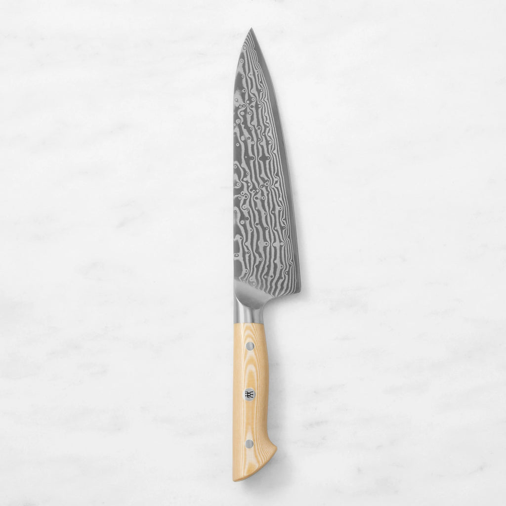 Damascus steel chef knife with a natural light wood handle and mosaic pin, centered vertically against a white marble background.