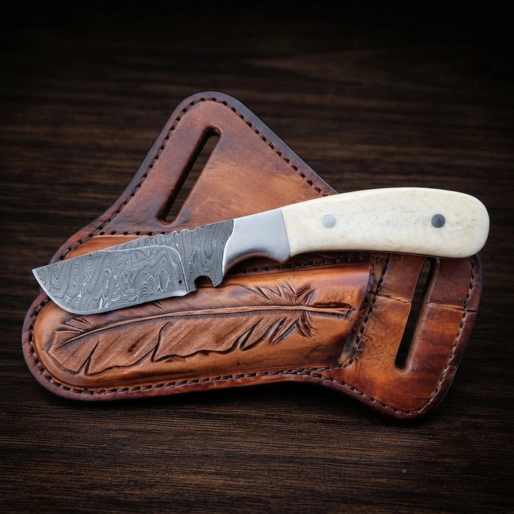 A handmade cowboy belt knife featuring a patterned Damascus steel blade with decorative filework on the spine, a polished steel bolster, and a smooth white bone handle with two silver pins, resting on top of a brown tooled leather pancake sheath carved with a feather design, on a dark wooden background