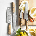 Handmade Damascus steel kitchen knives featuring light wood handles with mosaic pins, displayed flat lay next to a wooden cutting board with sliced citrus fruits.