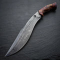 Handcrafted full tang Damascus steel Kukri knife featuring a contoured rosewood burl handle, resting diagonally on a dark slate stone surface