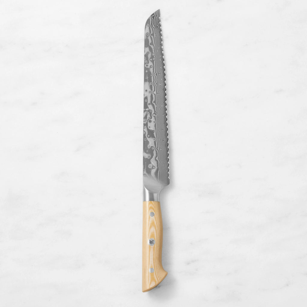 Damascus steel serrated bread knife with a natural light wood handle, centered vertically against a smooth white marble background