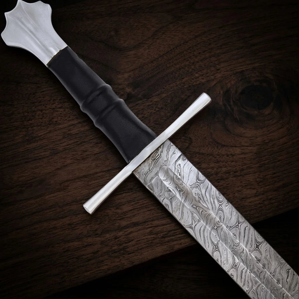 A hand-forged medieval sword resting diagonally on a dark wood background, highlighting the wave-like patterns of its Damascus steel blade, a polished straight crossguard, a contoured black leather grip, and a fluted pommel