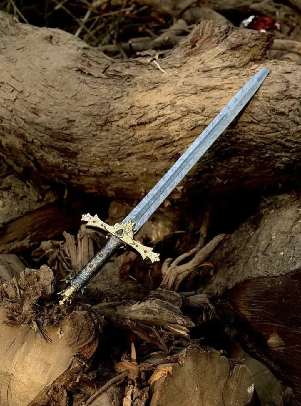 Handcrafted Damascus Steel sword with a wavy patterned blade resting on wooden logs, featuring a highly detailed gold and black ornate hilt with red gemstone accents.