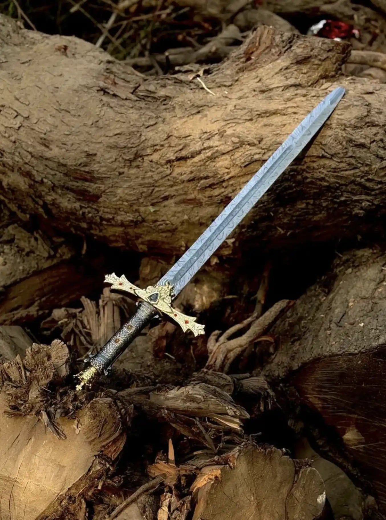 Handcrafted Damascus Steel sword with a wavy patterned blade resting on wooden logs, featuring a highly detailed gold and black ornate hilt with red gemstone accents.