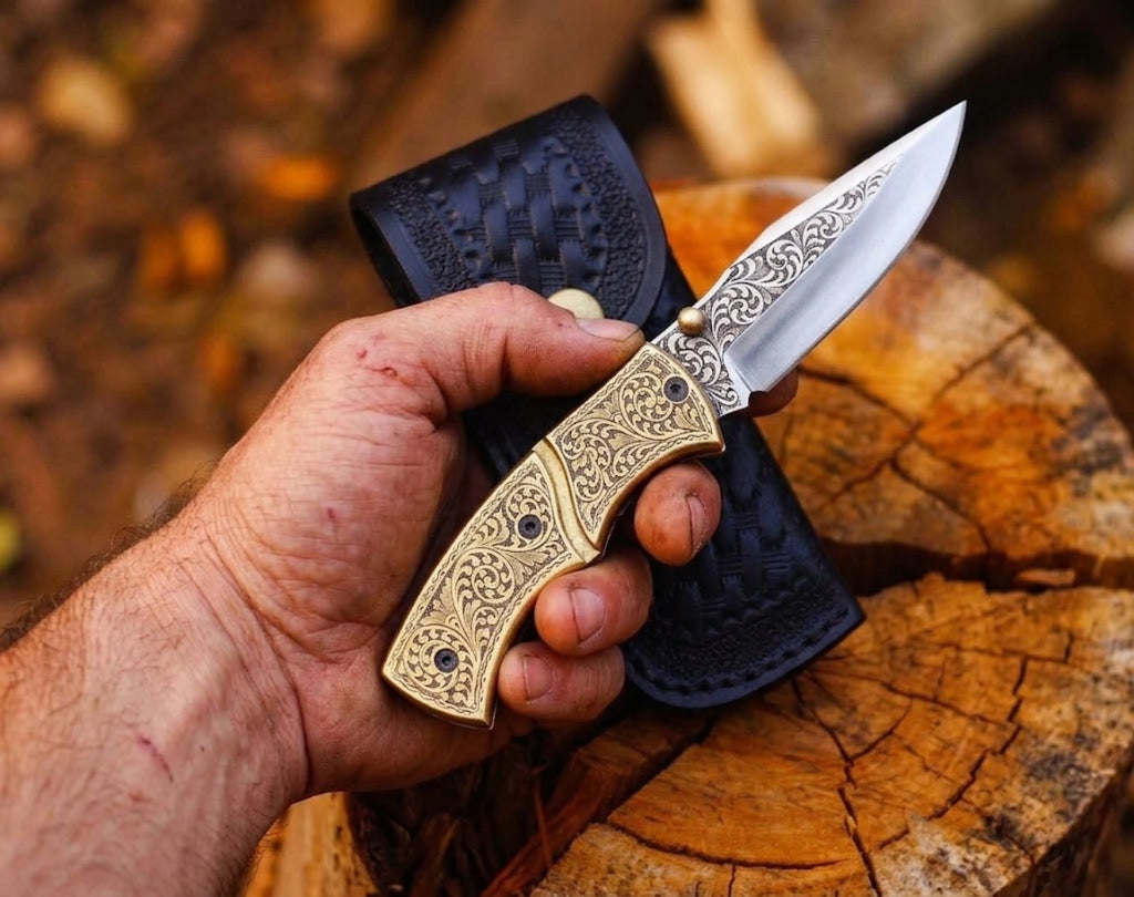 Hand holding a custom engraved brass folding pocket knife with detailed scrollwork on the handle and blade, held in front of a black leather sheath over a tree stump outdoors.