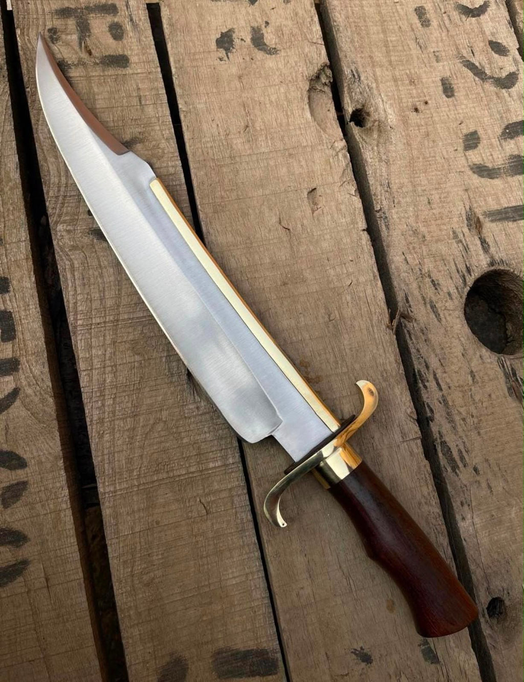 Full-length view of a handmade Bowie knife featuring a clip-point blade with a brass spine strip, polished brass double guard, and dark wooden handle on a wooden plank surface