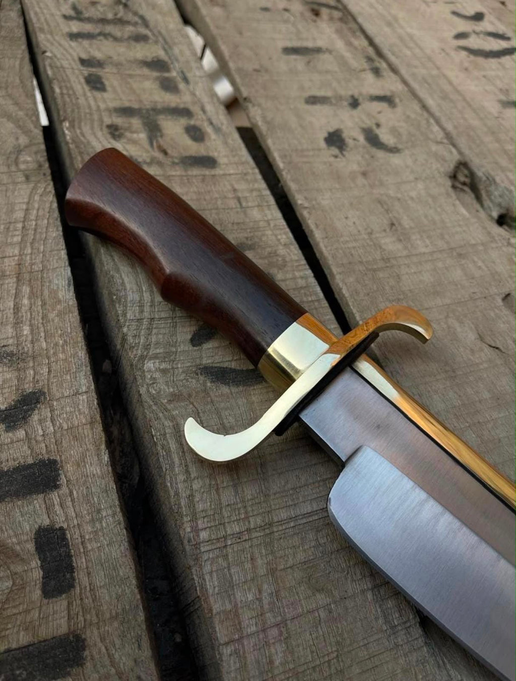 Angled close-up view of a handmade Bowie knife featuring an ergonomic dark wooden handle, polished brass double crossguard, and brass spine reinforcement on weathered wooden boards