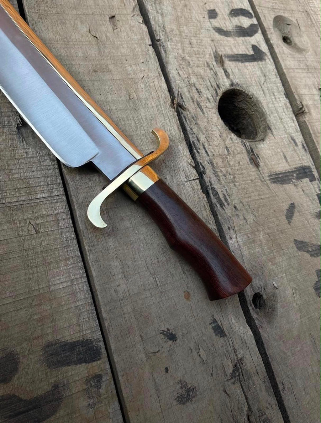 Close-up view of a handmade Bowie knife showing the curved brass double guard, dark wooden handle, and brass spine accent on a rustic wooden plank.