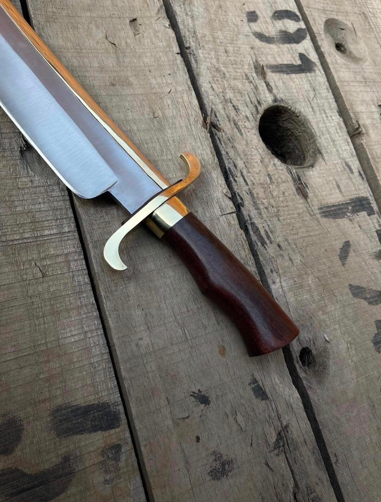Close-up view of a handmade Bowie knife showing the curved brass double guard, dark wooden handle, and brass spine accent on a rustic wooden plank.