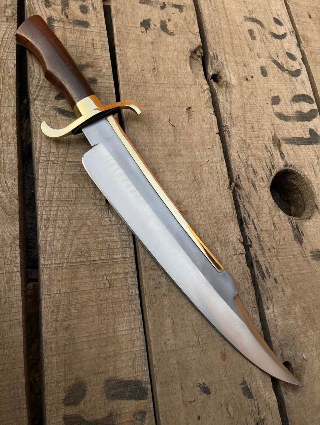 Full-length view of a handmade Bowie knife featuring a clip-point steel blade with brass spine lining, polished brass double guard, and rich wooden handle displayed on rustic wood planks.