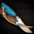 A custom handmade D2 tool steel knife with a highly polished mirror-finish blade and bolster, featuring a vibrant turquoise stone composite handle with dark veins, secured with silver pins, a lanyard hole with a tan leather strap, and a purple wood accent spacer, lying diagonally over a tan leather sheath on a dark wooden background.
