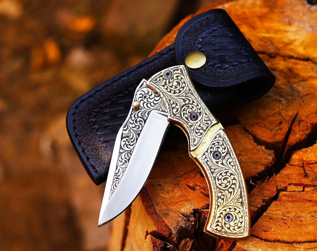 An angled view of a handmade engraved brass folding pocket knife, partially open, resting on a rustic wooden tree log in front of a black leather sheath