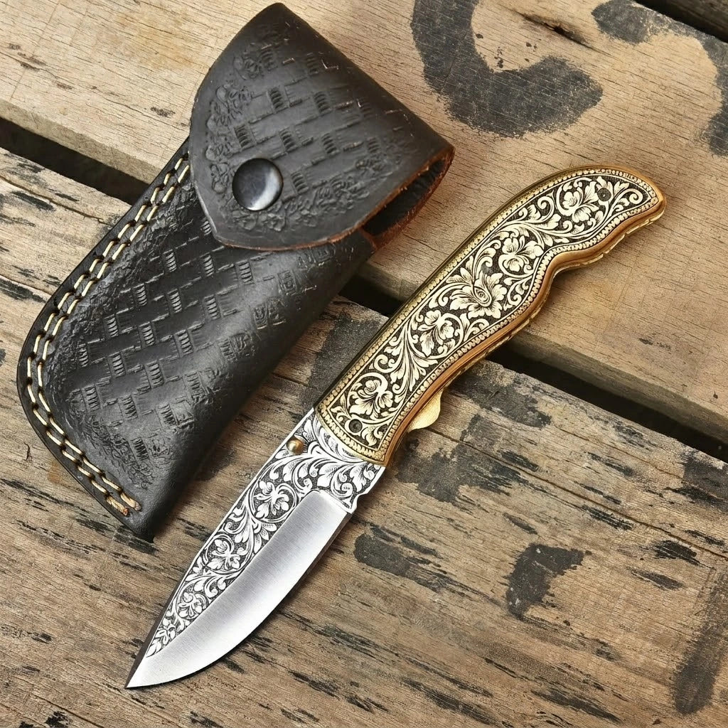 Handmade folding pocket knife featuring intricate floral engravings on the blade and brass handle, resting beside a dark brown stitched leather sheath on a rustic wooden surface