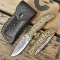 Handmade engraved brass folding pocket knives shown in both open and closed positions alongside a dark brown stitched leather sheath on a rustic wooden background.