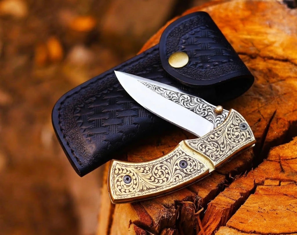 Handmade engraved brass folding pocket knife displayed partially open on top of a black leather sheath resting on a wooden tree stump