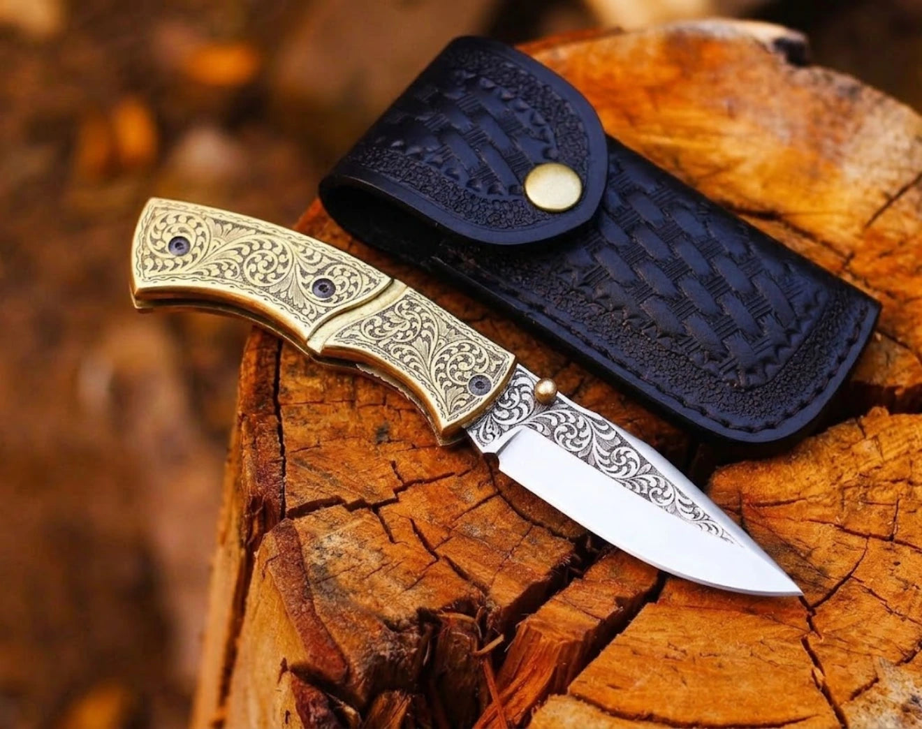 Handmade folding pocket knife with intricate scroll engraving on the brass handle and blade, resting on a weathered wooden tree stump beside a black leather sheath